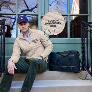 Peut inclure: Un homme porte un pull zipp&eacute; matelass&eacute; beige avec le texte "CUSTOM EMBROIDERY HERE". Il porte une casquette bleue et un pantalon vert. Un sac de sport noir est &agrave; c&ocirc;t&eacute; de lui sur les marches.