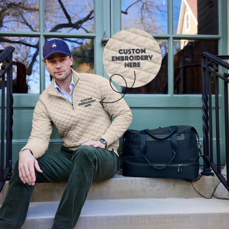 Peut inclure: Un homme porte un pull zipp&eacute; matelass&eacute; beige avec le texte "CUSTOM EMBROIDERY HERE". Il porte une casquette bleue et un pantalon vert. Un sac de sport noir est &agrave; c&ocirc;t&eacute; de lui sur les marches.