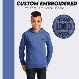 May include: A blue youth fleece hoodie with a pocket on the front. The hoodie has a black embroidered circle on the left chest with the text "YOUR LOGO HERE".