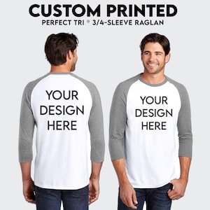 May include: A white and gray baseball t-shirt with 3/4 sleeves. The shirt has a white front and gray sleeves. The text "YOUR DESIGN HERE" is printed on the front and back of the shirt.