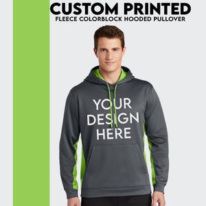 May include: A gray fleece hooded pullover with green colorblock panels on the sleeves. The hoodie has a drawstring hood and a kangaroo pocket. The text "YOUR DESIGN HERE" is printed on the front of the hoodie.