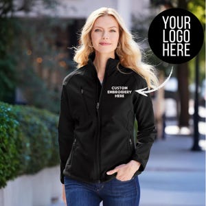 May include: A black softshell jacket with a full zipper, two side pockets, and a chest pocket. The jacket has the text "CUSTOM EMBROIDERY HERE" embroidered on the chest. A black circle with the text "YOUR LOGO HERE" is in the upper right corner.