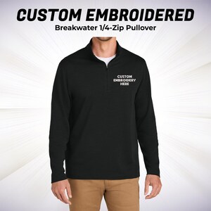 May include: Black Breakwater 1/4-Zip Pullover with the text "CUSTOM EMBROIDERED" at the top. The pullover has a white undershirt and the text "CUSTOM EMBROIDERY HERE" on the chest. The model is wearing khaki pants.