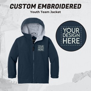 May include: A navy blue youth team jacket with a zipper closure and a hood. The jacket has a gray fleece lining and a patch on the left chest that says "YOUR DESIGN HERE".