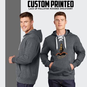 Puede incluir: Una sudadera con capucha de color gris oscuro con un detalle de encaje blanco en el cuello. La sudadera presenta un estampado gr&aacute;fico de un cohete amarillo con el texto "Startup" debajo.