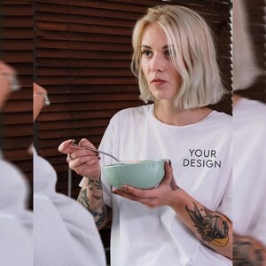 May include: A woman wearing a white t-shirt with the text "YOUR DESIGN" printed on it, is holding a light green bowl and a silver spoon. She has short blonde hair and tattoos on her arms.