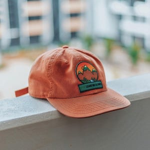 May include: A burnt orange baseball cap with a green, brown, and orange mountain scene patch and the text "Leave Me Alone" on the front.