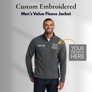 May include: A charcoal gray fleece jacket with a full zipper, featuring the text "YOUR TEXT" and "YOUR DESIGN HERE" in white. The jacket is part of a custom embroidered collection. The model is wearing dark blue jeans.