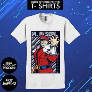 May include: White t-shirt with a colorful illustration of M. Bison from Street Fighter. The illustration shows M. Bison in his signature red and black outfit, with his signature pose and a determined expression. The text "M. Bison" and "Street Fighter" are printed on the shirt.