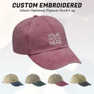 May include: A maroon custom embroidered Adams Optimum pigment dyed cap with a curved brim. The cap features a white square with the text "YOUR DESIGN HERE". Below are caps in olive, green, and maroon.