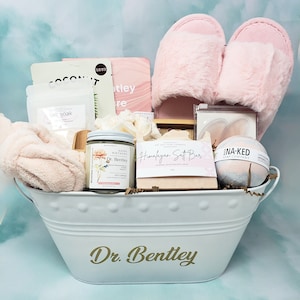 Peut inclure: Un panier en métal blanc rempli d'articles de soins personnels. Comprend des pantoufles roses duveteuses, une bougie, des bombes de bain, une barre de sel de l'Himalaya et des masques faciaux. Le panier porte l'inscription "Dr. Bentley" en lettres dorées.
