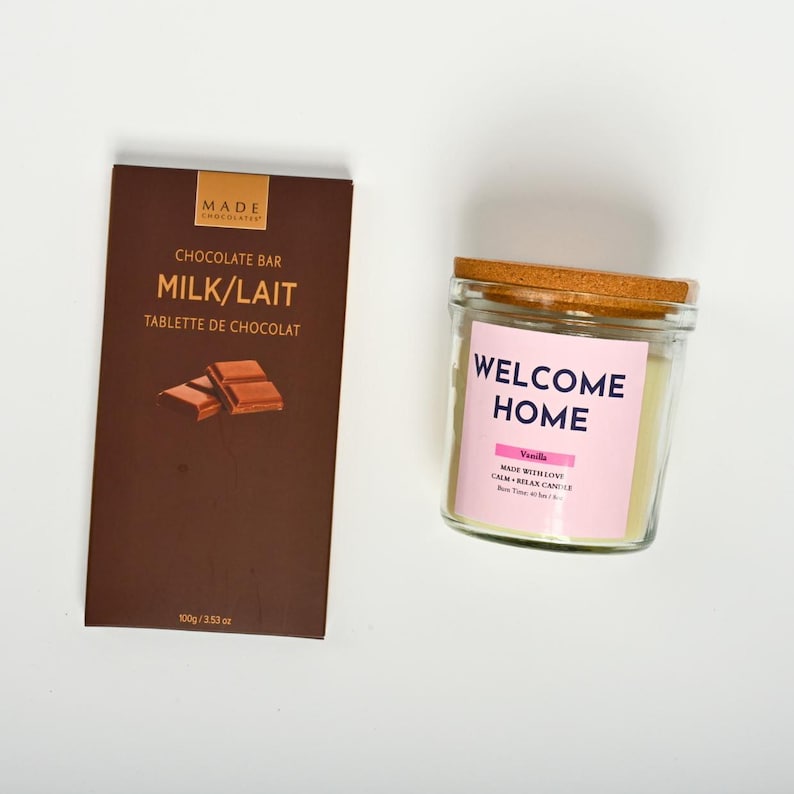Puede incluir: Una barra de chocolate marr&oacute;n con el texto "MADE CHOCOLATER" y "CHOCOLATE BAR MILK/LAIT TABLETTE DE CHOCOLAT" y una vela blanca con tapa de corcho y el texto "WELCOME HOME" en rosa y "Vanilla MADE WITH LOVE CALM + RELAX CANDLE BURN TIME 40-45 HRS" en negro.