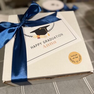 May include: A white gift box tied with a navy blue satin ribbon. A card on top reads "HAPPY GRADUATION Anna" with a graduation cap illustration. A small round sticker says "handmade with love".