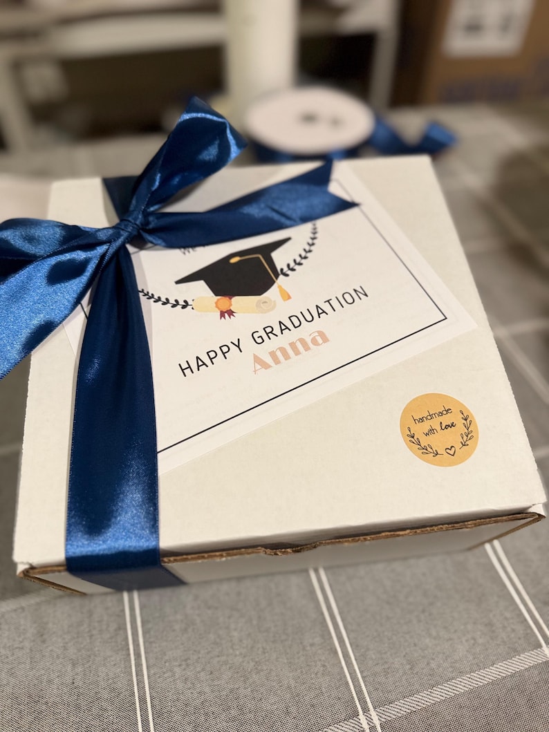 May include: A white gift box tied with a navy blue satin ribbon. A card on top reads "HAPPY GRADUATION Anna" with a graduation cap illustration. A small round sticker says "handmade with love".