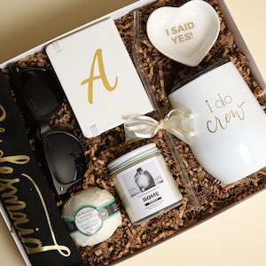 Könnte beinhalten: Eine weiße Geschenkbox gefüllt mit Artikeln für eine Brautjungfer. Die Box enthält eine schwarze Tragetasche mit goldener Aufschrift "bridesmaid", Sonnenbrillen, ein weißes Notizbuch mit einem goldenen Buchstaben "A", eine weiße Kerze mit einem Foto eines Paares, eine weiße Badebombe, einen weißen Becher mit goldener Aufschrift "i do crew", eine herzförmige Schale mit goldener Aufschrift "I said yes!" und einen Strohhalm.