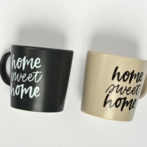 Puede incluir: Dos tazas de cer&aacute;mica con el texto "home sweet home" escrito en ellas. Una taza es negra con texto blanco y la otra es beige con texto negro.