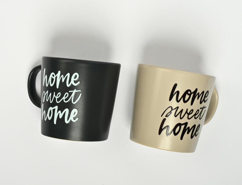 Puede incluir: Dos tazas de cer&aacute;mica con el texto "home sweet home" escrito en ellas. Una taza es negra con texto blanco y la otra es beige con texto negro.