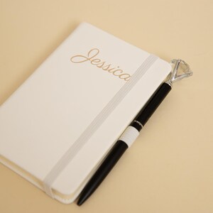 May include: Ivory-colored notebook with the name "Jessica" in gold script. A black pen with a diamond-shaped top rests beside the notebook. The notebook has a gray elastic band closure.