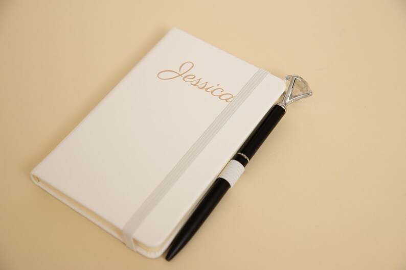 May include: Ivory-colored notebook with the name "Jessica" in gold script. A black pen with a diamond-shaped top rests beside the notebook. The notebook has a gray elastic band closure.