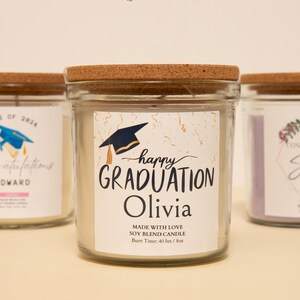 May include: Clear glass jar candle with a cork lid. The label reads "happy GRADUATION Olivia" with a graduation cap graphic. The candle is a soy blend and has a burn time of 40 hours. Other candles are visible in the background.