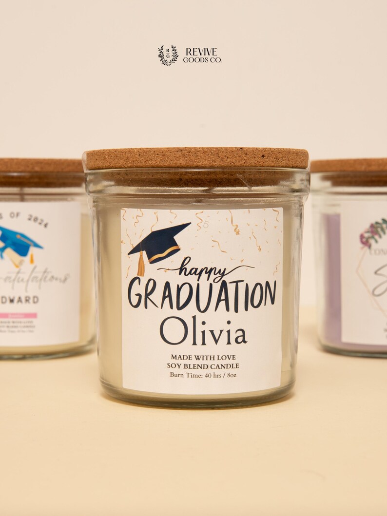 May include: Clear glass jar candle with a cork lid. The label reads "happy GRADUATION Olivia" with a graduation cap graphic. The candle is a soy blend and has a burn time of 40 hours. Other candles are visible in the background.