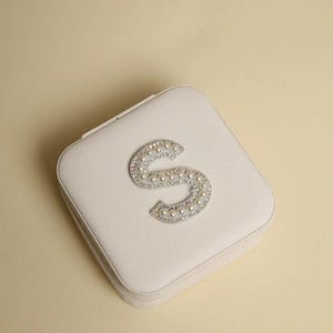 May include: A square, off-white jewelry box with a decorative letter "S" on the lid. The letter is embellished with small, shiny, silver-colored rhinestones and faux pearls. The box is closed and has a zipper.