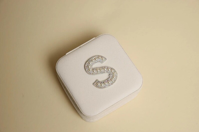 May include: A square, off-white jewelry box with a decorative letter "S" on the lid. The letter is embellished with small, shiny, silver-colored rhinestones and faux pearls. The box is closed and has a zipper.