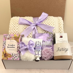 Peut inclure: Un panier-cadeau blanc rempli de produits de bain et de corps à la lavande, une couverture blanche, une boîte de truffes au cacao et une tasse personnalisée avec le nom "Ashley".