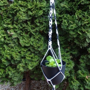 Op de afbeelding: Een zwarte macramé plantenhouder met witte en marineblauwe accenten, met een zwarte pot met groene planten. De houder hangt aan een zwarte metalen stang tegen een achtergrond van groen gebladerte. Het macramé ontwerp bevat een kwast.