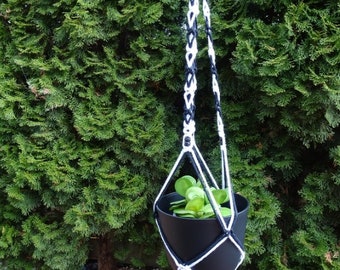 May include: A black macrame plant hanger with white and navy blue accents, holding a black pot with green plants. The hanger is suspended from a black metal bar against a backdrop of green foliage. The macrame design includes a tassel.