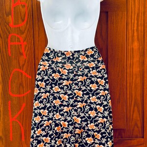 May include: A black skirt with an orange floral pattern is displayed on a white mannequin. The skirt features a repeating design of orange flowers and white vines against a black background. The skirt is knee-length.