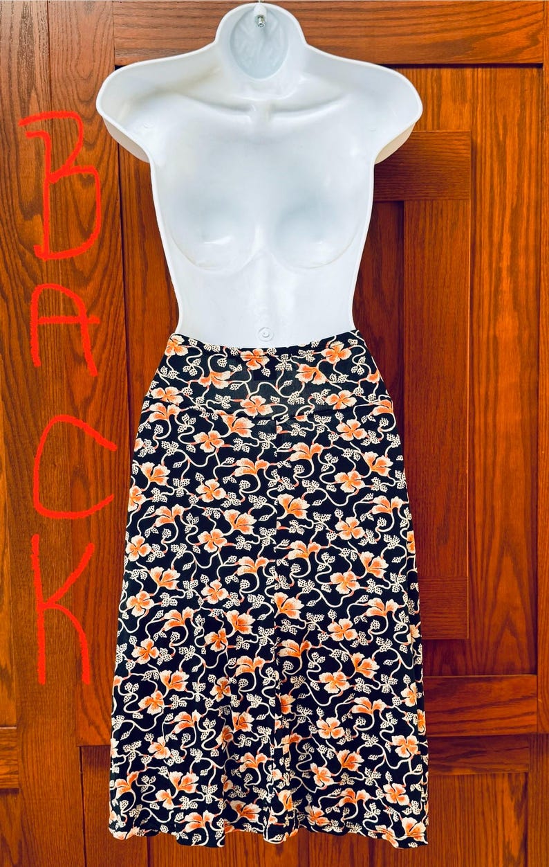 May include: A black skirt with an orange floral pattern is displayed on a white mannequin. The skirt features a repeating design of orange flowers and white vines against a black background. The skirt is knee-length.