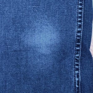 May include: Close-up of blue denim fabric with a lighter blue faded area. The image shows the texture of the denim and a vertical seam with detailed stitching on the right side. The overall tone is a classic, worn look.