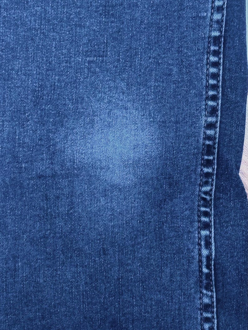 May include: Close-up of blue denim fabric with a lighter blue faded area. The image shows the texture of the denim and a vertical seam with detailed stitching on the right side. The overall tone is a classic, worn look.