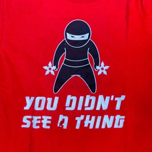 May include: Red t-shirt with a black ninja cartoon holding throwing stars and the text "YOU DIDN'T SEE A THING" in white.