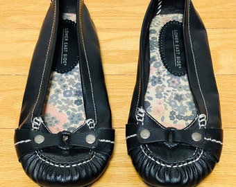 May include: A pair of black leather ballet flats with a white stitch detail and a bow on the toe. The shoes have a floral print insole and say "Lower East Side" on the inside.