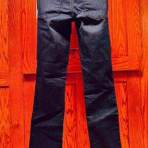 May include: Dark wash denim jeans with a classic five-pocket design. The jeans have a straight leg cut and are displayed against a wooden background. The back pockets and belt loops are visible.