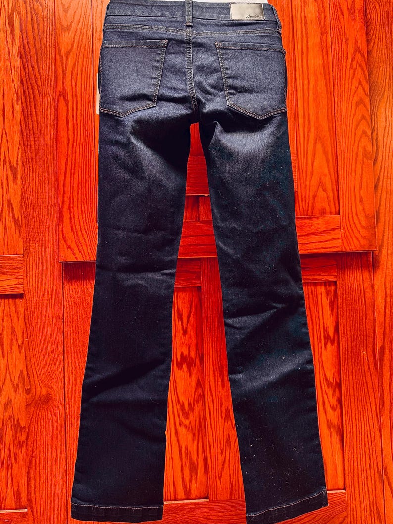 May include: Dark wash denim jeans with a classic five-pocket design. The jeans have a straight leg cut and are displayed against a wooden background. The back pockets and belt loops are visible.