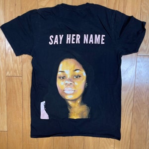 May include: Black t-shirt with a portrait of a woman and the text "SAY HER NAME" printed on the back.