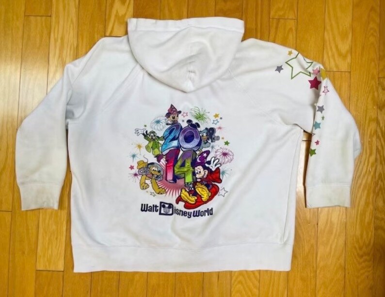 K&ouml;nnte beinhalten: Wei&szlig;er Kapuzenpullover mit einem bunten Aufdruck von Mickey Mouse und seinen Freunden, die 2014 in Walt Disney World feiern. Der Pullover hat Sterne auf den &Auml;rmeln.