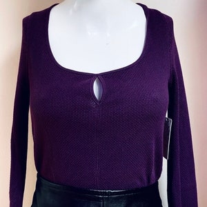 May include: A long-sleeved, purple waffle knit top with a keyhole cutout at the neckline. The top is worn with a black leather skirt.