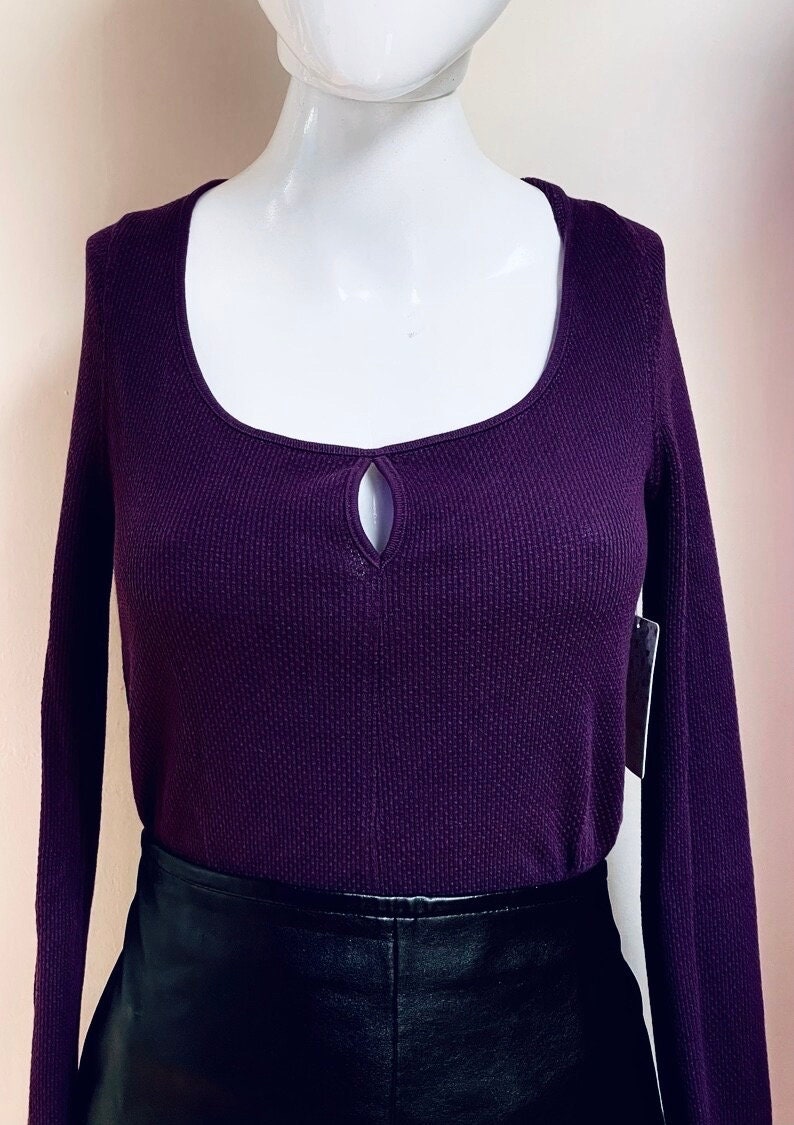 May include: A long-sleeved, purple waffle knit top with a keyhole cutout at the neckline. The top is worn with a black leather skirt.