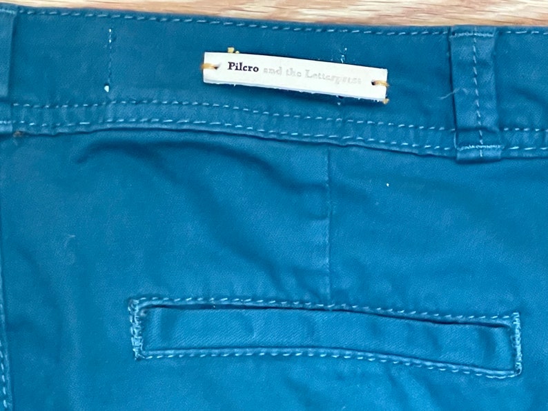 Puede incluir: Primer plano de pantalones de color verde azulado con un bolsillo trasero y una etiqueta que dice "Pilero and the Letterpress". Los pantalones tienen costuras visibles y un dise&ntilde;o cl&aacute;sico.