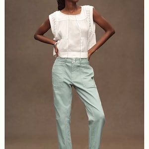 Puede incluir: Una mujer modela unos vaqueros relajados de talle medio en color verde claro. Los vaqueros tienen el bajo vuelto y se combinan con una camiseta blanca y sandalias negras. La camiseta tiene un escote cuadrado y un patr&oacute;n de rejilla.