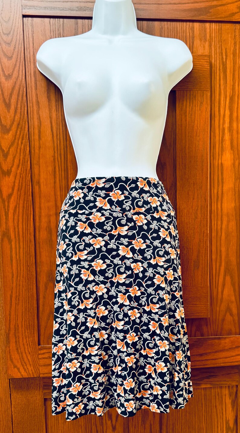 May include: A black skirt with a floral pattern in orange and white, displayed on a white mannequin. The skirt has a high waist and a flowing design, perfect for a casual or semi-formal look.