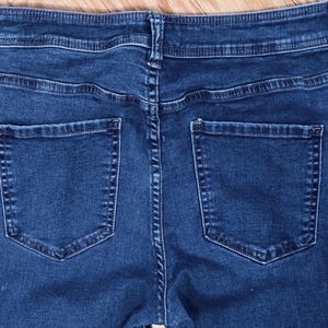 May include: Close-up of the back of dark blue denim jeans. The jeans have two back pockets and a waistband with belt loops. The stitching is visible, and the fabric appears to be a medium weight. The jeans are laid flat on a wooden surface.