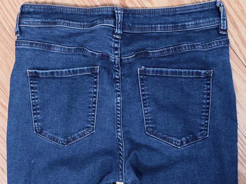 May include: Close-up of the back of dark blue denim jeans. The jeans have two back pockets and a waistband with belt loops. The stitching is visible, and the fabric appears to be a medium weight. The jeans are laid flat on a wooden surface.