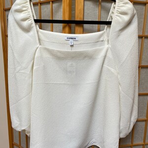 May include: White, long-sleeved blouse with a square neckline and puffed sleeves. The blouse has a textured fabric and a label that says "EXPRESS".