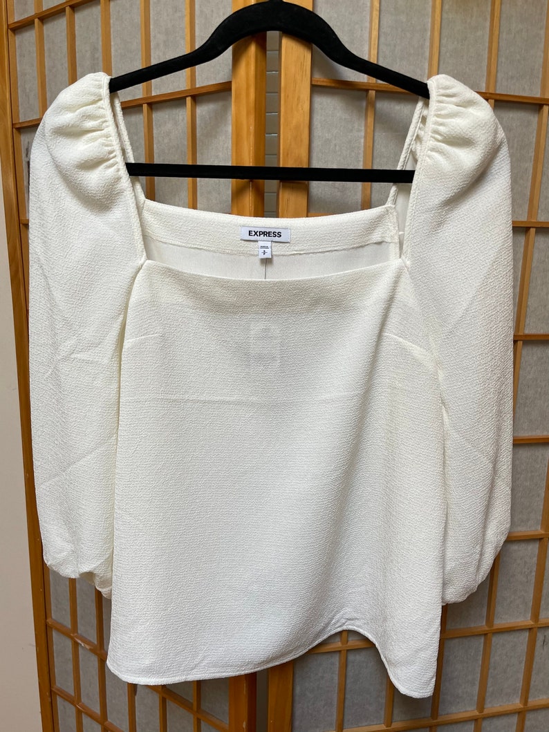 May include: White, long-sleeved blouse with a square neckline and puffed sleeves. The blouse has a textured fabric and a label that says "EXPRESS".