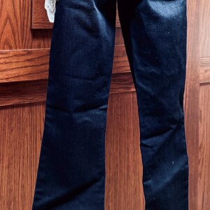May include: Dark wash denim jeans with a classic five-pocket design. The jeans have a straight leg and a slight fade. A tag is attached to the jeans. The jeans are displayed against a wooden background.